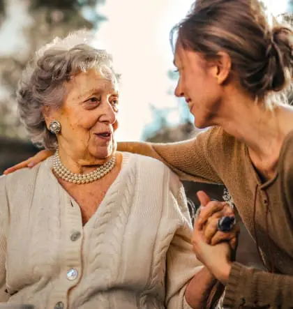 A younger woman holds the hand of an older woman and smiles at her.
