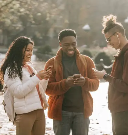 Three young people stand together outside looking at their phones and smiling.