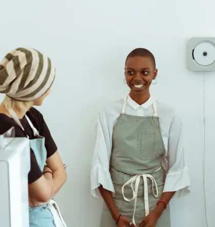 A person in an apron standing against a wall smiles at another person in a hat an apron standing in profile in front of them.