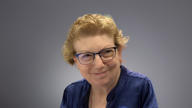 An image of Leslie Carpenter, a white woman with glasses and short hair and a blue shirt smiling at the camera over her shoulder.